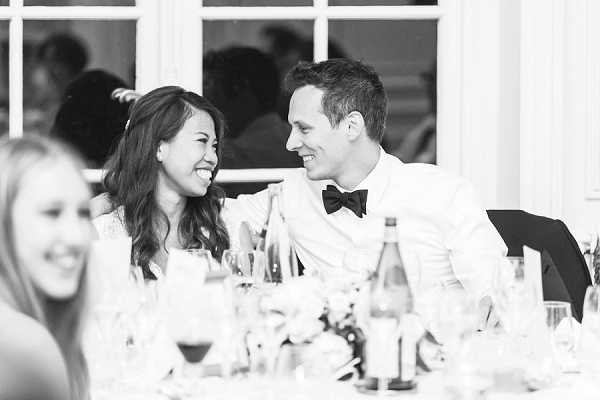 Black-and-white close-up portrait of a bride and groom seated at the head table during an indoor wedding reception, laughing and looking at each other. The groom wears a white dress shirt with a black bow tie, and the bride has shoulder-length wavy hair with a small hair accessory. The table in the foreground is set with glassware, wine bottles, and floral centerpieces. A blurred female guest is visible in the left foreground, and additional guests appear softly out of focus in the background. The image has bright highlights and soft mid-tones, with large white-framed windows visible behind the couple.
