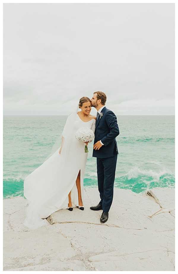 A couple portrait taken outdoors on a flat coastal rock directly at the water's edge, with the ocean visible behind them. The bride wears a long-sleeved white gown with a front slit, a cathedral-length veil blowing in the wind, and black two-tone cap-toe shoes; she holds a compact bouquet of white baby's breath with small pink accents. The groom wears a navy suit with a patterned navy tie and a small floral boutonniere, and is kissing the bride on the temple as she smiles. The image is a full-length portrait shot in natural light with an overcast sky.