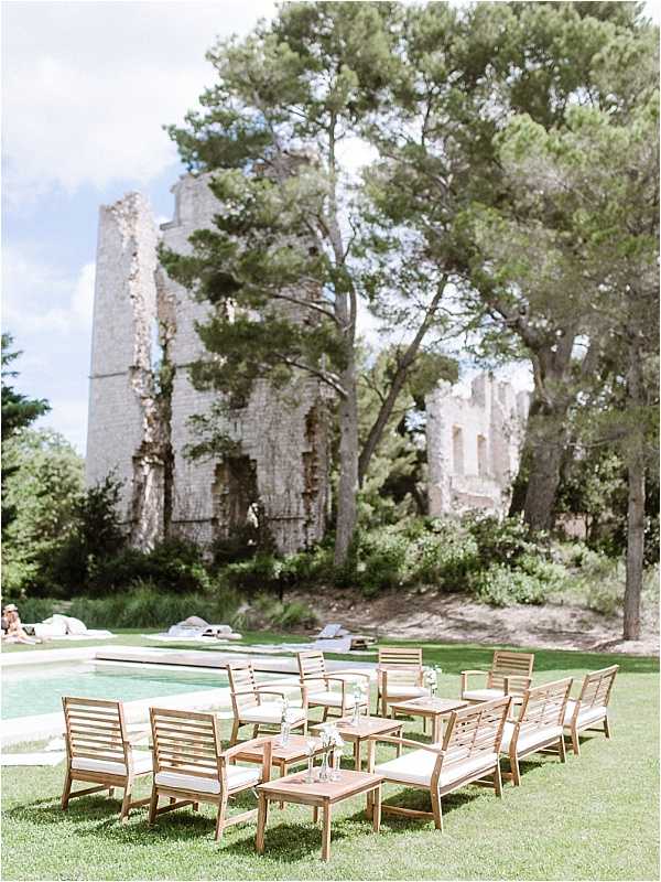 An outdoor cocktail hour or lounge seating area set up on a lawn beside a pool, with large stone ruins of what appears to be a historic building or abbey rising in the background. The seating arrangement consists of approximately ten teak wood armchairs with cream or off-white cushions, grouped around several small square teak side tables, each holding small bud vases with delicate white flowers. A few guests can be seen relaxing near the pool in the background. The styling is clean and understated, pairing natural wood tones with neutral upholstery against the dramatic backdrop of the weathered stone ruins. Wide shot composition. Potential venue feature image.