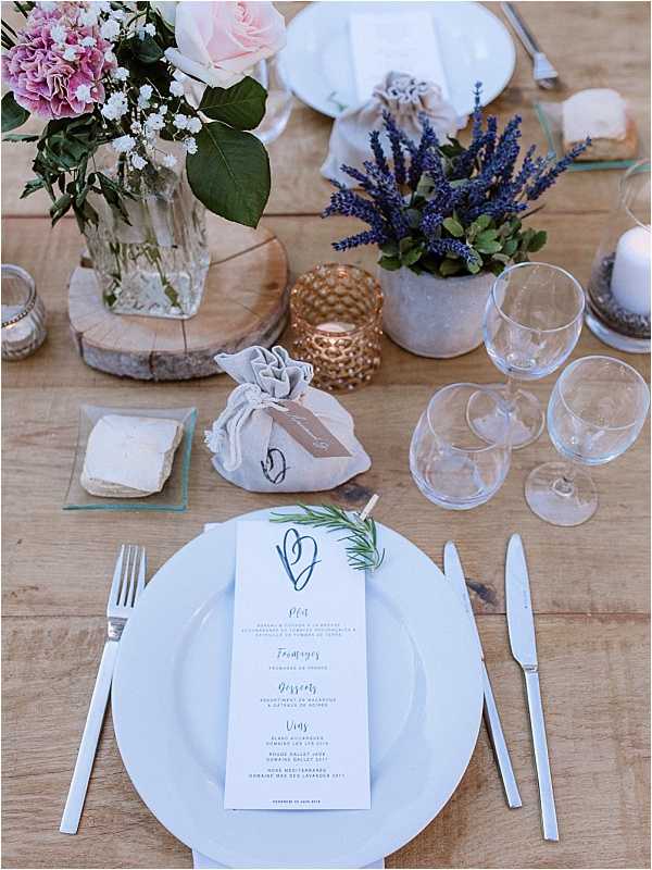 Close-up detail shot of a wedding reception place setting on a bare wooden farm table. The setting includes a white charger plate with a printed menu card featuring a hand-drawn heart motif and a sprig of fresh rosemary clipped to it, flanked by silver flatware. A small linen drawstring favor bag with a kraft tag sits beside the plate. Centerpiece elements visible include a wood slice riser holding a glass vase with blush pink roses, pink carnations, and green foliage, a small concrete pot with dried blue lavender, a gold hammered metal tealight votive, a white pillar candle, and two empty wine glasses. The overall styling theme is rustic-Provençal with a palette of blush pink, blue-purple lavender, gold, and white accents.