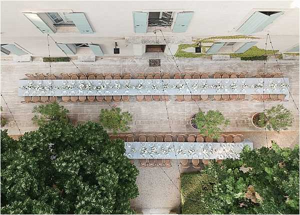 Aerial shot of an outdoor wedding reception setup in a stone courtyard, likely at a French chateau or historic estate. Three long banquet tables are arranged in parallel rows, dressed with pale blue-grey linen runners and topped with white and pale green floral centerpieces spaced evenly along their length. Tan/cognac-colored rattan or wicker chairs line both sides of each table, accommodating a large number of guests. String lights are strung overhead across the courtyard, and potted trees and mature green foliage frame the space. The building facade visible at the top of the frame features pale turquoise-blue shuttered windows. Potential venue feature image.