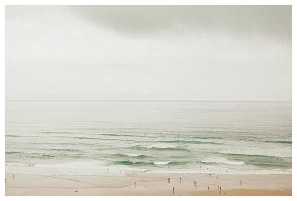 This image does not appear to be a wedding photograph. It is a wide aerial or elevated landscape shot of a beach with pale sand, gentle surf with soft green-tinted waves, and an overcast grey sky. Several small figures are visible on the shoreline but no wedding activity, couple, bridal party, or wedding-related decor is present. The image is not suitable for a wedding content library as it contains no identifiable wedding elements.