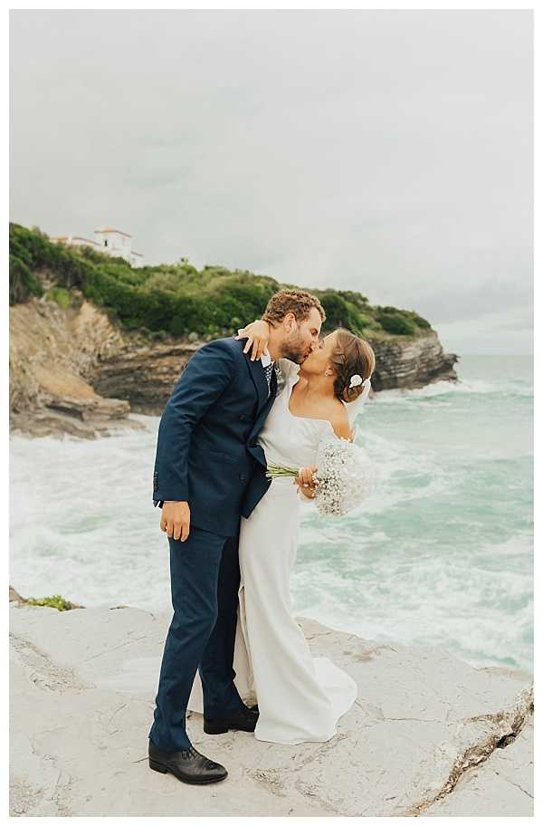 A couple shares a kiss during outdoor wedding portraits on a rocky coastal clifftop, with crashing waves and a headland visible in the background. The groom wears a navy blue suit with black dress shoes and a patterned tie, while the bride wears a long-sleeve, off-the-shoulder white fitted gown and holds a small bouquet of baby's breath. A white flower is tucked into the bride's updo hairstyle. The shot is a full-length portrait with a natural, relaxed coastal setting and an overcast sky, giving the image a muted, moody color palette consistent with a boho-coastal styling aesthetic.