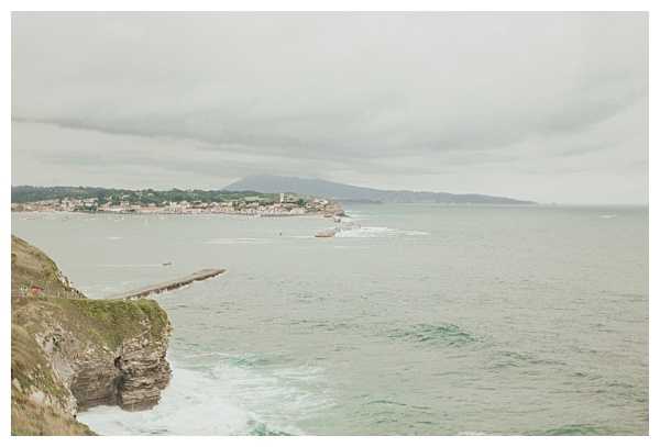 Wide landscape shot taken from a clifftop overlooking a coastal bay under an overcast sky. No people, wedding couple, or wedding-related decor are visible in this image. The scene shows a small coastal town in the distance, a jetty or pier extending into the water, and cliffs in the foreground. This appears to be a location/venue establishing shot rather than a wedding photograph with subjects.