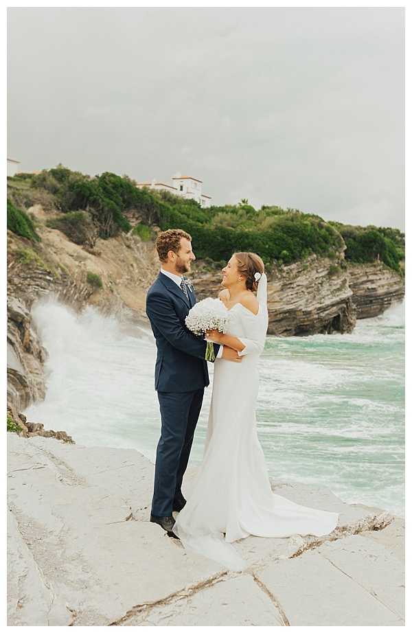 A couple portrait taken outdoors on a rocky coastal clifftop, with waves crashing dramatically behind them. The groom wears a navy blue suit with a patterned tie, and the bride wears an off-the-shoulder ivory fitted gown with a long train and a short white veil; she holds a small round bouquet of white baby's breath. The two face each other and smile, embracing lightly. The setting appears to be the Basque coastline near Biarritz, with a white villa visible on the cliffside in the background. The composition is a full-length portrait shot with a slightly overcast, moody atmosphere.