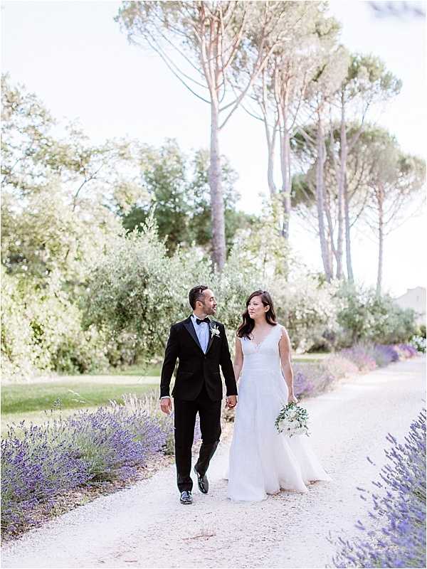A couple portrait taken outdoors on a gravel pathway lined with purple lavender on both sides. The bride wears a white A-line gown with a V-neckline and carries a round bouquet of white flowers with greenery, while the groom wears a black tuxedo with a bow tie and a small white boutonniere. The two are walking hand-in-hand along the path, looking at each other, with tall umbrella pines and silver-leafed shrubs visible in the background. The image is a medium full-length shot with soft, bright natural light giving it an airy, light-toned quality consistent with a film or film-inspired style.
