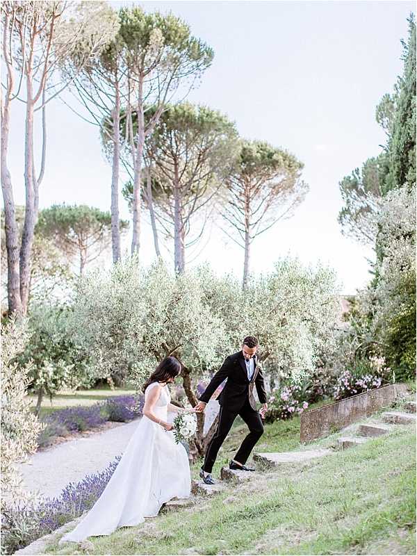 The bride and groom are navigating stone garden steps together in an outdoor portrait shot, with the groom helping guide the bride down carefully. The setting is a formal garden landscape featuring tall umbrella pine trees, olive trees, and flowering shrubs. The bride wears a white sleeveless ball gown with a full skirt and carries a white and green bouquet, while the groom is dressed in a black tuxedo with a bow tie. The composition is a wide environmental portrait with soft, even natural lighting and a classic, clean styling aesthetic.