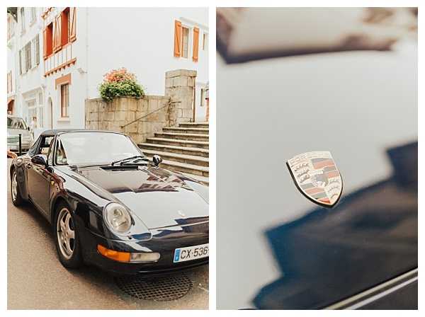 A two-image composite showing a dark navy blue Porsche 911 (993 generation) parked on a street in front of a white stone building with orange shutters and exterior steps, used as a wedding car. The left panel is a wide shot capturing the full vehicle from a front three-quarter angle, while the right panel is a close-up detail shot of the Porsche crest badge on the hood, showing the gold, red, and black enamel emblem. Both images are shot in a warm, film-style color tone. No people are visible; the focus is entirely on the vehicle as a styled wedding detail.