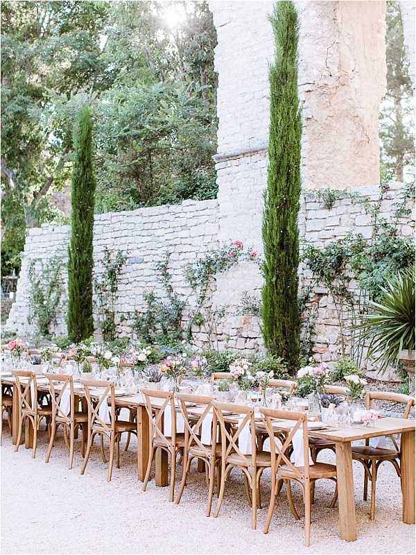 An outdoor wedding reception table setup in a walled garden or courtyard, styled in a relaxed rustic-classic aesthetic. Two long wooden farm tables run parallel, lined with natural oak cross-back chairs and dressed with white linen napkins, glassware, and bud vases containing blush pink, coral, and cream florals including what appear to be roses and ranunculus scattered along the center. The setting features a gravel floor and a tall stone wall backdrop framing the scene, with tall columnar cypress trees rising between the architectural stone pillars. Wide establishing shot capturing the full table arrangement and surrounding outdoor environment. Potential venue feature image.