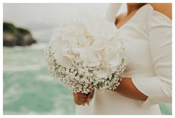 Close-up detail shot of a bride holding a rounded bridal bouquet composed of white hydrangeas surrounded by baby's breath, photographed against a coastal backdrop with turquoise-green water visible in the background. The bride is wearing a long-sleeved white satin dress with a clean, minimalist silhouette. The bouquet has a compact, dome-shaped arrangement with the baby's breath forming a delicate frilled border around the central hydrangea blooms. The overall styling is clean and minimal, with an all-white floral palette.
