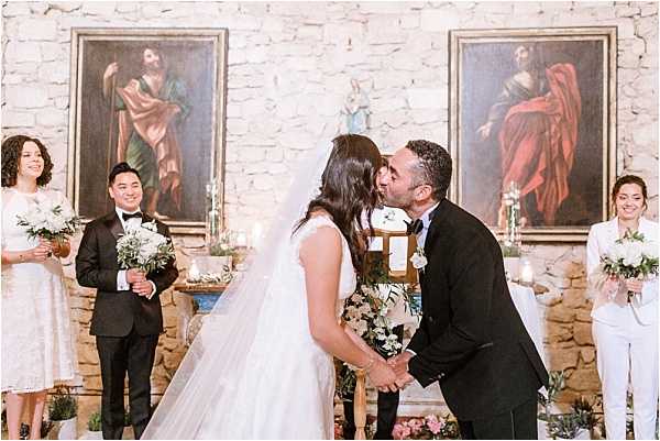 The bride and groom share their first kiss during an indoor ceremony in a small chapel or historic stone-walled room. The bride wears a white ballgown with a long veil, and the groom is dressed in a black tuxedo. Two witnesses stand on either side of the couple — a man in a black suit holding a small white and greenery bouquet on the left, and a woman in a white trouser suit holding a bouquet of white flowers on the right. The altar behind them is decorated with candles and small floral arrangements, and two large oil paintings depicting religious figures hang on the exposed stone wall. The ceremony setting has a classic, intimate feel with warm candlelight. Wide portrait-style composition.