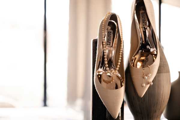 Close-up detail shot of a pair of cream-colored Badgley Mischka pointed-toe heels displayed upright on a dark surface, photographed against a bright window background. The shoes feature gold metallic heel hardware and are accessorized with a delicate pearl strand bracelet draped inside one shoe and a small gold drop earring with a pearl accent resting against the other. The overall color palette is cream, gold, and ivory, suggesting a classic bridal styling aesthetic. The image is softly backlit with a shallow depth of field, giving the foreground shoe sharp focus while the background is gently blurred.