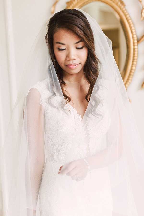 A bridal portrait of a single bride photographed indoors, likely during a getting-ready moment. She wears an ivory lace gown with long sheer sleeves and a deep V-neckline, paired with a long cathedral-style veil draped over her head and shoulders. Her dark hair is styled in loose waves swept to one side, and her makeup features neutral tones with defined eyes. A gold ornate mirror is partially visible on the wall behind her. The composition is a close-up portrait with soft, bright natural light creating an airy, light-filled atmosphere.
