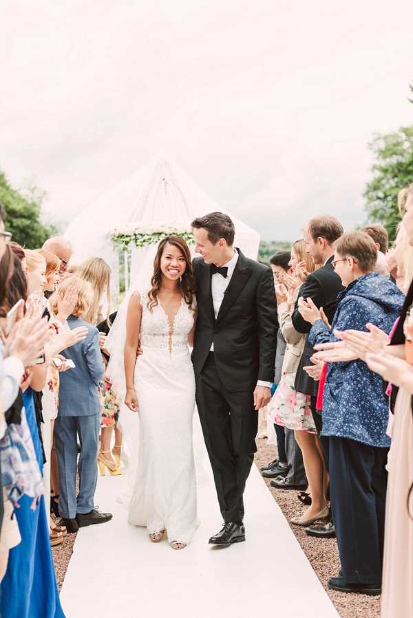 A newly married couple walks back down the aisle during their outdoor ceremony recessional, surrounded by approximately 20 applauding guests on either side of a white aisle runner. The bride wears a fitted lace gown with a deep V-neckline, sheer embroidered bodice, and a long veil, paired with strappy heeled sandals; the groom wears a black tuxedo with a black bow tie. In the background, a white tent or canopy structure with a floral greenery arch is visible, indicating the ceremony took place outdoors. The couple is smiling and looking at each other, while guests in colorful summer attire clap and cheer in a wide, candid portrait-style shot.