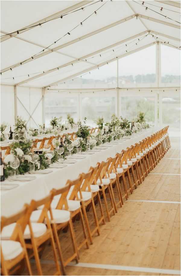 A reception setup inside a white marquee tent with clear glass panel walls and a string of bistro lights running along the ceiling. The image shows a single long banquet table dressed in a white linen tablecloth, lined on both sides with natural wood folding chairs with white padded seats. The table centerpiece runs the full length with a lush greenery garland interspersed with white florals, likely white roses and eucalyptus, along with clear glassware, gray cloth napkins, and dark wine bottles placed at intervals. The overall decor palette is white and green with natural wood tones, giving a clean, modern-rustic aesthetic. Wide shot taken from one end of the table, emphasizing the length of the setup and the light-filled marquee interior.