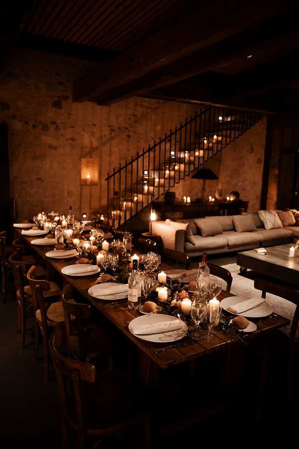 A wedding reception dinner table setup photographed in a wide shot inside a rustic stone-walled venue with exposed wooden beam ceilings and an iron staircase railing. The long dark wood farm table is set for approximately 14 guests, with white plates, folded white napkins, multiple wine glasses per setting, and scattered pillar and votive candles providing warm, low ambient lighting. The table runner features dark foliage, small dried or neutral-toned floral elements, and wine bottles placed along the center. Bentwood chairs line both sides of the table. In the background, a lounge area with a grey sofa and additional candles lining the staircase steps creates a moody, intimate atmosphere consistent with a rustic or industrial-chic styling theme.