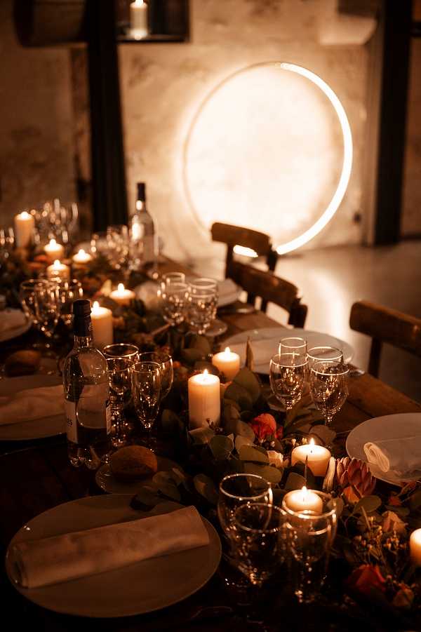 A detail shot of a wedding reception tablescape in a dimly lit indoor venue with rustic stone walls. The long wooden dining table is styled with a greenery and mixed floral runner featuring terracotta and rust-toned blooms, scattered pillar candles in varying heights, crystal glassware including wine glasses and champagne flutes, white ceramic plates with folded linen napkins, and wine bottles placed along the table. The dominant background feature is a large circular neon halo light mounted on a textured plaster wall, casting a warm amber glow across the entire scene. The overall styling theme is moody and modern-rustic, with the candlelight and neon circle creating a warm, low-light atmosphere throughout the frame.