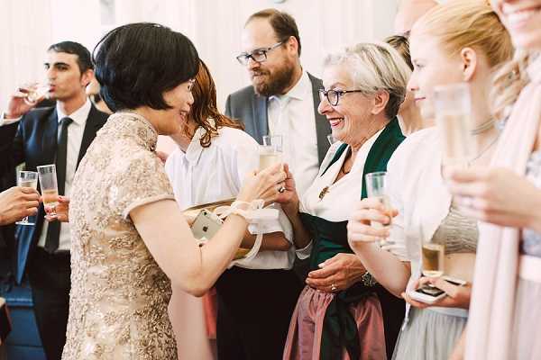 A cocktail hour or drinks reception scene captured in a candid, mid-shot composition showing approximately six guests mingling indoors. At the center of the frame, a woman in a gold lace short-sleeve dress holds a champagne flute and appears to be in warm conversation with an older woman wearing a traditional dirndl-style outfit with a dark green bodice and dusty rose skirt. Several other guests are visible holding champagne glasses, including a man in a dark suit in the background and a woman in a blush dress on the right edge of the frame. The setting appears to be a bright, light-filled interior space. The gathering has a relaxed, celebratory atmosphere with a mix of formal and traditional attire among the guests.