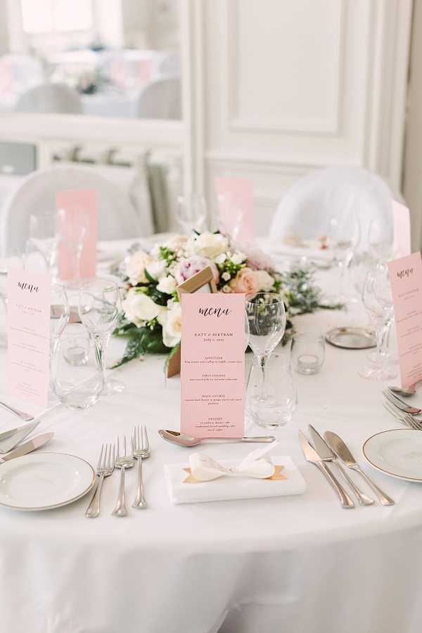 A close-up detail shot of a wedding reception table set in an indoor ballroom or salon with white paneled walls visible in the background. The round table is dressed in a white linen with white china, silver cutlery, and crystal stemware at each place setting. Blush pink printed menu cards reading 'menu' in script are placed at each setting, personalized with the couple's names and a July date. The centerpiece is a low, lush floral arrangement of ivory roses, blush pink peonies, and greenery with what appears to be a gold geometric vessel. A white folded napkin with a small gold detail sits at the center place setting. The overall decor palette is blush pink, white, and gold, giving the tablescape a classic, romantic aesthetic.