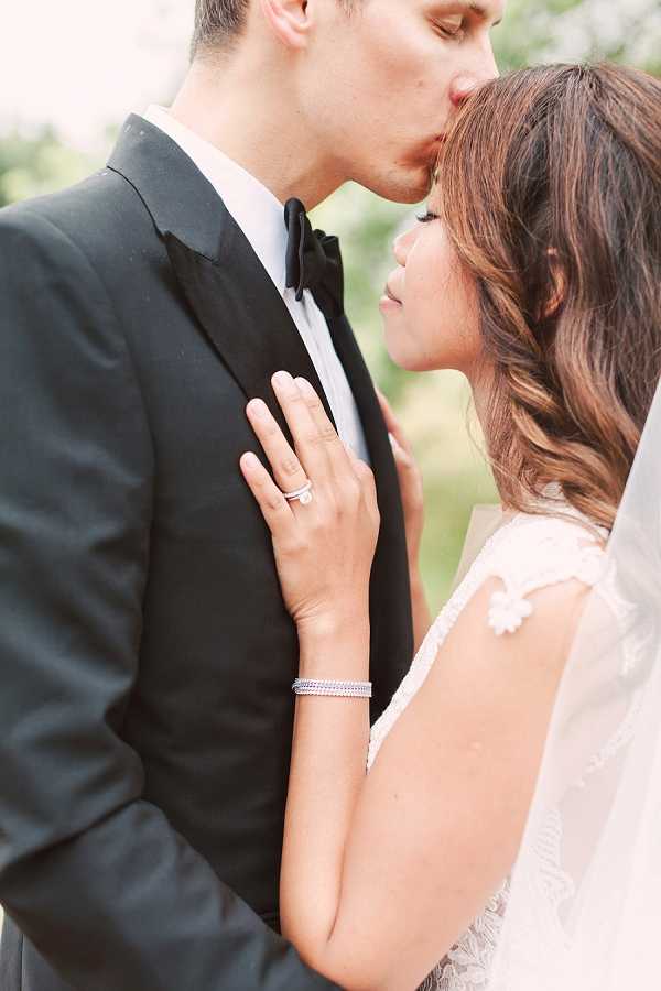 A close-up portrait of a bride and groom sharing a kiss outdoors, with a softly blurred green background. The groom wears a dark charcoal suit with a white dress shirt and black bow tie, while the bride wears a white lace dress with cap sleeves and a flowing veil visible at the right edge of the frame. The bride's hand rests on the groom's chest, showcasing a diamond solitaire engagement ring with a wedding band and a delicate crystal or diamond bracelet on her wrist. Her dark brown hair is styled in loose waves. The image is shot in a tight, intimate portrait composition with shallow depth of field, emphasizing the couple's faces and the bride's jewelry details.