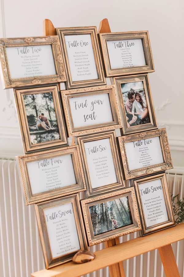 A wedding seating chart display consisting of multiple ornate gold-toned picture frames arranged in a collage style on a wooden easel. The frames contain calligraphy table assignment cards labeled Table One through Table Seven, a central 'Find your seat' sign in script lettering, and several engagement or couple photos interspersed throughout. The table cards list guest names in handwritten-style script on white backgrounds. The frames feature decorative embossed detailing in an antique gold finish, and the overall decor palette is warm gold and ivory. A small touch of greenery is visible at the lower right. The display is photographed against a white interior wall with a white radiator, suggesting an indoor venue space. This is a medium-distance detail shot capturing the full easel arrangement.