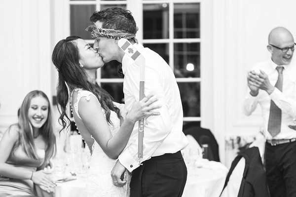 A black-and-white close-up portrait of a bride and groom sharing a kiss on the dance floor during what appears to be their first dance at an indoor reception. The bride wears a sleeveless lace dress and the groom is in a white dress shirt with suspenders and dark trousers, with what appears to be a small Canadian flag sticker visible on his cheek and a decorative headband or mask pushed up on his forehead, suggesting a playful or game-related moment. In the background, a female guest laughs and a male guest in a tie and glasses applauds, with reception tables set with glassware visible. The image has soft contrast with bright highlights from large white-framed windows behind the couple.