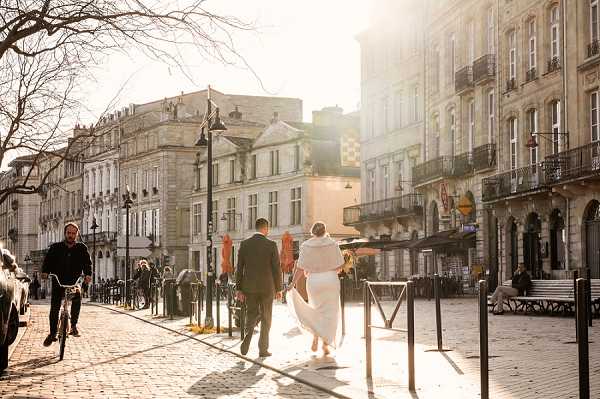 A bride and groom are walking together through a cobblestone city square or plaza, shot from behind in a wide environmental portrait. The setting is an urban French city center with classic Haussmann-style limestone buildings lining the street. The bride wears a white gown with a white fur or faux-fur wrap stole, and the groom is in a dark suit. Strong golden backlighting creates a warm sun flare effect across the scene. Several passersby are visible, including a man on a bicycle to the left, giving the image a candid, documentary feel. The composition is a wide shot that emphasizes the urban surroundings as much as the couple.
