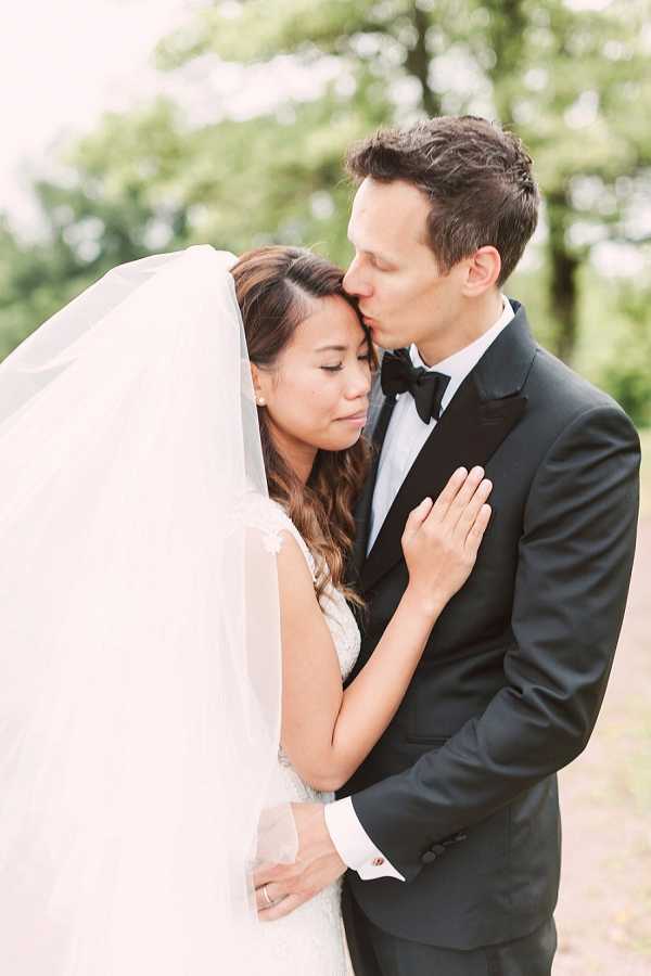 An outdoor couple portrait showing the groom kissing the bride on her temple while she rests her hands on his chest with eyes closed. The bride wears a white lace gown with a long flowing veil, and the groom is dressed in a black tuxedo with a black bow tie and white dress shirt with cufflinks. The composition is a mid-shot with soft, diffused natural light and a blurred green foliage background, giving the image a light, airy feel with a fine-art film aesthetic.