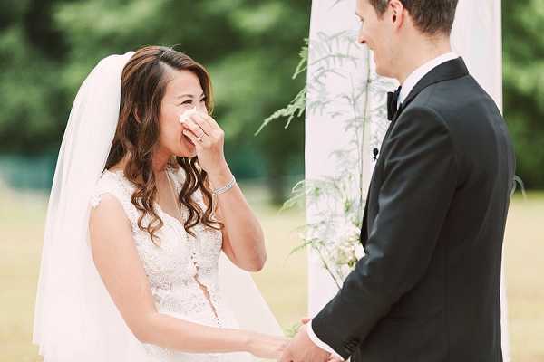 An outdoor wedding ceremony capturing an emotional moment between the couple during vow exchange, with the bride visibly crying and pressing a tissue to her nose while holding the groom's hand. The bride wears a white lace cap-sleeve gown with a cathedral veil and a delicate bracelet, with her dark hair styled in loose waves. The groom is dressed in a classic black tuxedo with a bow tie, shown in profile. Behind them is a white draped ceremony arch decorated with thin green foliage, set against an outdoor garden backdrop. This is a close-up portrait shot with a shallow depth of field.