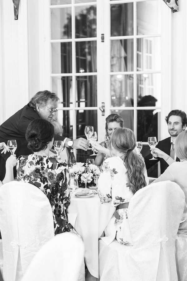 Black-and-white image of a wedding reception toast at a round dining table, with approximately six guests raising wine glasses. An older man in a dark suit leans in to clink glasses with those seated, including two women in floral-patterned dresses and a younger man in a suit. The chairs are covered in white slipcovers, and a low floral centerpiece is visible on the table. The setting appears to be an indoor reception room with tall white French doors in the background, suggesting a classic or formal venue. The image is a medium shot taken from a slightly elevated angle, with bright highlights and soft mid-tones typical of high-key black-and-white photography.