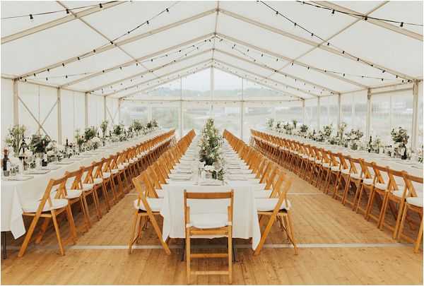 Wide shot of a fully set wedding reception inside a clear-sided white marquee tent with a peaked roof, showing three long banquet-style tables arranged in parallel rows with white linen tablecloths and natural wood folding chairs. The decor palette is white and green, with low centerpieces of greenery and white florals spaced along each table, accompanied by glassware, white napkins, and wine bottles. String lights are strung across the interior roofline, adding warm overhead lighting. The light-wood flooring and transparent marquee walls allow natural daylight to fill the space, giving it a clean, airy, modern-rustic aesthetic.