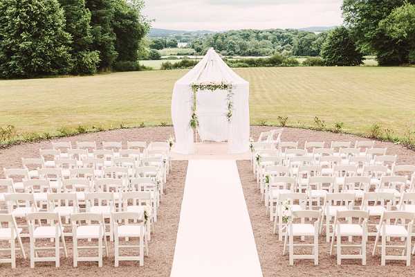 An outdoor wedding ceremony setup photographed from a slightly elevated wide-angle perspective, showing a symmetrical arrangement of white folding chairs divided into two sections by a white aisle runner. At the far end of the aisle stands a square arch decorated with greenery and small white florals, framed by a draped white fabric canopy structure. The ceremony space is set on a gravel or packed earth circular area, and no guests or wedding party are present, suggesting the photo was taken before the ceremony began. The overall decor palette is white and green, with a classic and clean styling aesthetic. Potential venue feature image.
