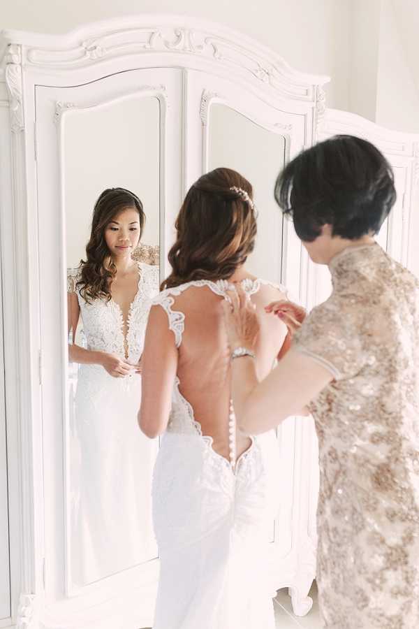 A bride is getting ready indoors, standing in front of a large ornate white tri-panel mirror while a second woman — wearing a gold and champagne sequined qipao-style top — fastens the back of the bridal gown. The bride wears a fitted ivory lace wedding dress with a deep V-neckline, sheer lace cap sleeves, a low open back, and a row of small buttons running down the spine. Her dark hair is styled in a soft half-up updo accessorized with a delicate crystal hair piece. The composition uses the mirror to simultaneously show the bride's front and back, and the bright, airy white room and ornate carved white armoire create a classic, light-filled bridal preparation scene.