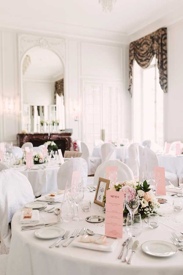 Indoor wedding reception setup in a classic French ballroom or chateau salon featuring white boiserie paneling, an ornate arched mirror above a dark marble fireplace, and tall windows with draped valance curtains. Multiple round tables are dressed in white floor-length linens with white chair covers, set with full silver cutlery, wine glasses, and white porcelain plates. The foreground table centerpiece features a low arrangement of blush pink peonies, white roses, and small white filler flowers with greenery, accompanied by a gold-framed table number card reading 'Table 6' and a blush pink printed menu card. The overall decor palette is white and blush pink with classic, formal styling. Wide shot capturing the full room layout with approximately six to eight tables visible. Potential venue feature image.