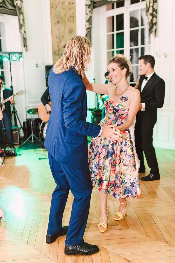 Two guests dance together on a parquet wood floor during the wedding reception in what appears to be an indoor ballroom or salon with ornate white paneling and large French windows. The man wears a cobalt blue suit with black shoes, and the woman wears a strapless midi-length dress with a multicolored floral print in pink, yellow, purple, and green, paired with yellow strappy heeled sandals. A live band performs in the background, with a guitarist visible, and another guest in a black tuxedo stands nearby. The setting has a classic French interior style, and the shot is taken at mid-range, capturing the dancers in full from a slight side angle.