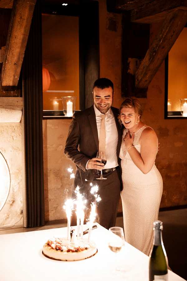 A couple stands together at a cake table during an indoor wedding reception, reacting with laughter and delight as sparkler candles ignite on a round cake decorated with small fruits. The groom wears a charcoal grey suit with a blush tie and holds a glass of red wine, while the bride wears a fitted sleeveless ivory lace gown with a V-neckline. The setting is a rustic interior space with exposed wooden ceiling beams, warm amber-toned walls, and candlelight visible on shelves in the background. A champagne bottle and glasses are placed on the white-clothed table beside the cake. The portrait-style shot is taken at medium distance with warm, low ambient lighting giving the scene a golden tone.