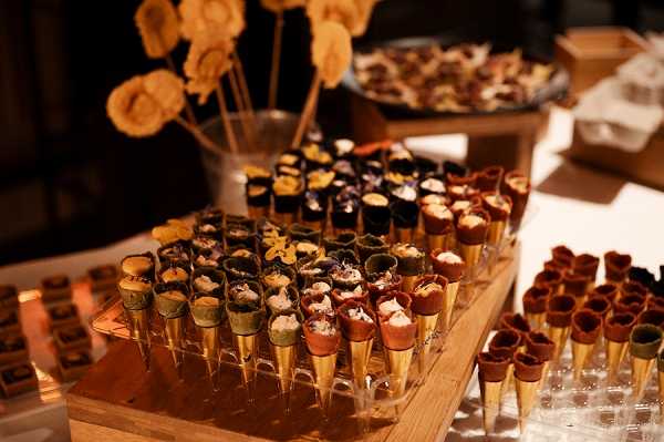 Close-up detail shot of a catering display at what appears to be a wedding cocktail hour or reception. A wooden tiered tray holds dozens of miniature savory or sweet cones in varied colors — dark chocolate, terracotta red, green, and natural — each filled with small toppings or fillings including creamy and colorful garnishes. Additional mini cones are arranged on a separate tray to the right, and a glass vessel holding thin wafer lollipops on skewers is visible in the background alongside a platter of other canapés. The display is set on a white-clothed table and lit with warm ambient lighting, suggesting an indoor evening reception setting. The warm golden tones of the wooden board and cone bases create a cohesive rustic-modern catering aesthetic.