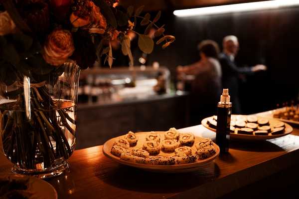 A close-up detail shot of a cocktail hour food station set up indoors, featuring a wooden board with bite-sized rolled canapés garnished with black sesame seeds, alongside a dark glass bottle and additional appetizers on a second wooden board in the background. A clear glass vase holds a loose arrangement of coral and orange garden roses with eucalyptus foliage, placed on a warm-toned wooden surface. The room is dimly lit with warm amber lighting, creating a moody atmosphere, and two guests — a man in a dark suit and a woman — are visible but out of focus in the background near what appears to be a bar area. The overall decor palette is dark and warm, with a modern, intimate styling approach.
