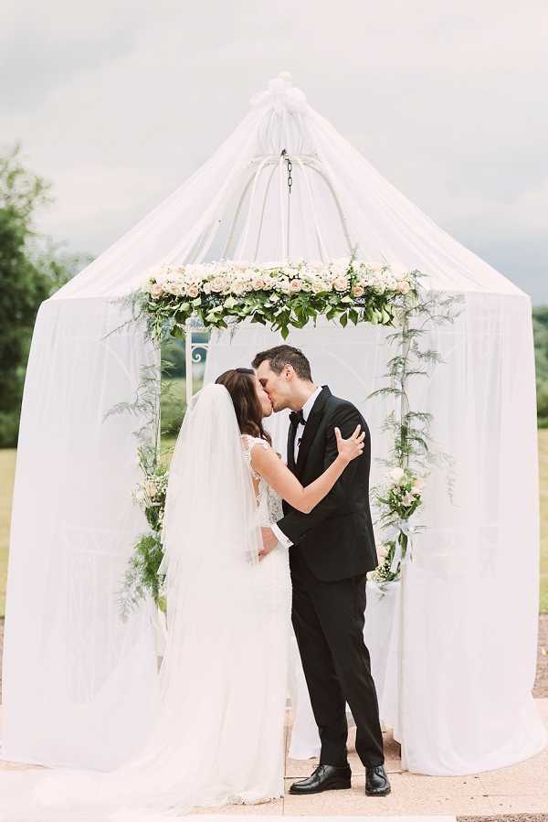 A bride and groom share a kiss during an outdoor ceremony beneath a white draped gazebo structure with a tent-like canopy. The gazebo is decorated with a floral garland of blush and ivory roses with lush greenery along its upper interior frame, and additional greenery and small florals accent the side posts. The bride wears a lace-detailed white gown with a long cathedral-length veil, while the groom wears a classic black tuxedo with a bow tie. The portrait is taken as a medium-wide shot with the couple centered against the white draped backdrop, set within an open outdoor garden or estate grounds setting with a classic, formal styling aesthetic.