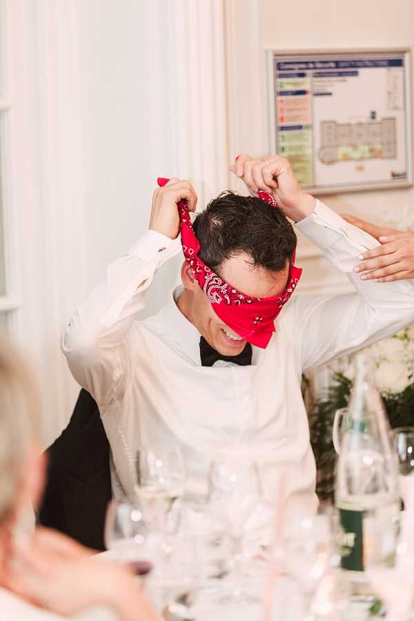 A candid reception moment captured at an indoor wedding dinner, showing a man in a white dress shirt and black bow tie being blindfolded with a red paisley bandana, likely as part of a wedding game. Another person's hands are visible helping to tie the bandana around his eyes, and he is smiling. The table in the foreground is set with wine glasses, water bottles, and white floral centerpieces. The room has white draped walls and a framed notice board is visible on the wall in the background. Medium portrait shot.