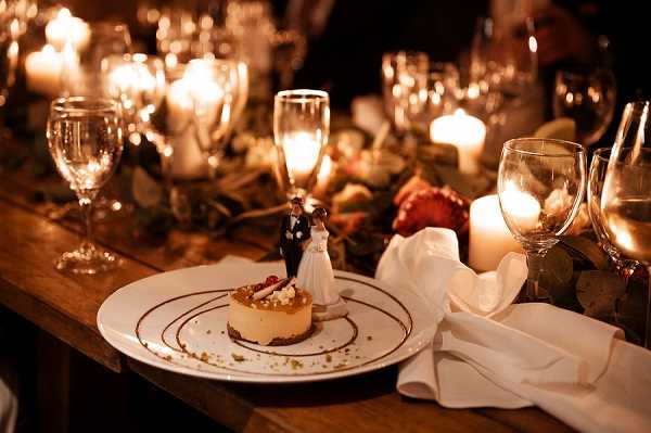 Close-up detail shot of a wedding reception table setting, showing a plated individual cheesecake dessert garnished with a chocolate sauce spiral, pistachio crumble, and a small classic bride-and-groom cake topper figurine placed on top. A white linen napkin is folded beside the plate, accompanied by wine glasses and a champagne flute. The table is lit entirely by candlelight from pillar candles and votive candles, creating a warm amber glow across the scene. The centerpiece runner includes greenery and what appears to be dark red florals, and the table itself is a rustic wooden farm-style surface, suggesting a warm, candlelit indoor reception with a rustic-classic styling.