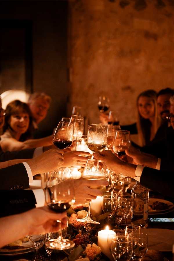 A toast during an indoor wedding reception dinner, with approximately eight guests raising glasses of red and white wine toward the center of the frame. The setting appears to be a stone-walled venue, likely a historic French building, lit almost entirely by candlelight — pillar candles and small votive candles run along the length of the table, casting a deep amber glow across the scene. The table is dressed with scattered glassware, plates with food, and a low floral arrangement featuring orange and rust-toned blooms. Guests in the background are dressed in dark formal attire. The composition is a close-up portrait-style shot focused on the clinking glasses in the foreground, with guests softly blurred behind, emphasizing the warm, candlelit atmosphere of the dinner.