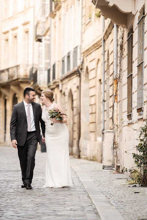 A couple portrait taken outdoors on a cobblestone street lined with classic French limestone buildings featuring shuttered windows and wrought-iron balconies. The bride wears a fitted, floor-length ivory lace gown and carries a loose bouquet featuring dusty pink and coral blooms with eucalyptus and mixed greenery. The groom is dressed in a charcoal grey suit with a pale pink tie. The two are walking hand-in-hand and smiling at each other. The shot is a medium-length portrait with a shallow depth of field, with the architecture softly blurred in the background, giving the urban street scene a warm, golden tone.