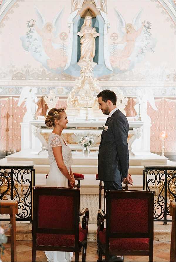 A bride and groom stand facing each other and holding hands at the altar during an indoor church ceremony. The bride wears a white gown with a lace cap-sleeve bodice and has her hair in an updo adorned with a small gold hair accessory, while the groom wears a dark navy textured suit with a white boutonniere. They are positioned in front of ornate dark wood and red velvet ceremonial chairs, with a decorative white altar behind them featuring a painted mural of angels in soft blue and pink tones and a central statue of the Virgin Mary. A small arrangement of pale flowers sits on the altar. The composition is a medium portrait shot taken from slightly behind and between the two chairs.