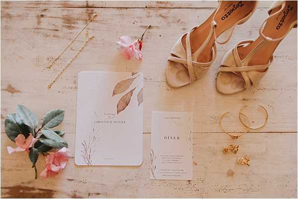 A flat lay detail shot of wedding stationery and accessories arranged on a whitewashed wooden surface. The stationery suite includes a white wedding invitation card with dried botanical leaf illustrations for 'Caroline & Olivier' and a matching white dinner menu card, both featuring a minimalist design with fine typography. Surrounding the stationery are nude/champagne strappy heeled sandals branded Repetto, a gold delicate chain bracelet, a gold cuff bracelet, small gold floral stud earrings, a gold ring, and two small pink roses with green leaves used as decorative accents.