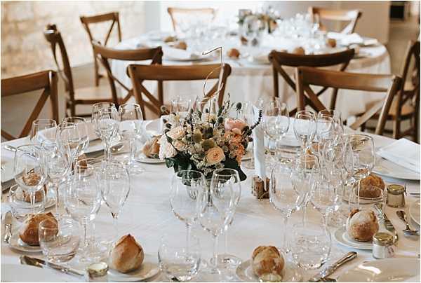 A close-up detail shot of a round reception table set for a wedding dinner inside what appears to be a stone-walled venue. The table is dressed with a white linen cloth and set with multiple wine and water glasses per place setting, white plates, silver cutlery, and individual bread rolls on side plates. A low centerpiece of blush and peach roses, cream stock flowers, and trailing greenery sits at the center, accompanied by a wire table number. Cross-back wooden chairs surround the table, and additional set tables are visible in the soft background, suggesting a classic French rustic reception style.