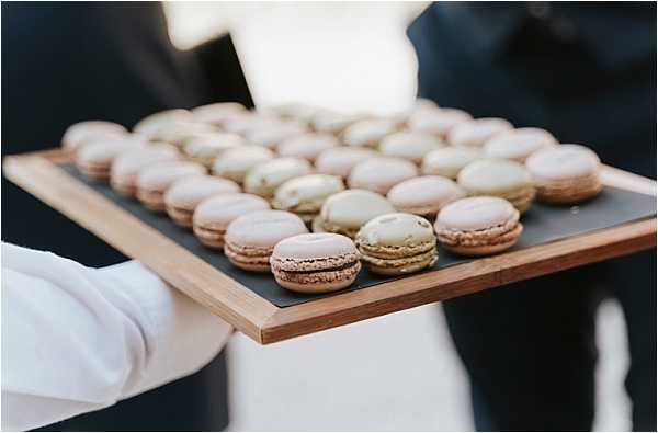 A close-up detail shot of a server in white gloves holding a wooden and slate tray filled with approximately 30 French macarons in two colors: pale blush-pink and sage green. The macarons are neatly arranged in rows on the tray, suggesting a cocktail hour or reception canapé service. A second person in a dark suit is partially visible in the background, slightly out of focus.