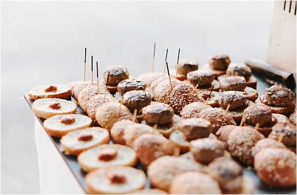 Close-up detail shot of a wedding catering spread featuring two rows of bite-sized food items arranged on a dark tray. On the left are open-faced mini brioches topped with what appears to be a red jam or chutney, and on the right are small sesame-seeded slider buns assembled into mini burgers, each secured with a cocktail pick. The tray is set on a white-clothed surface, consistent with a cocktail hour food station. The background is softly blurred and neutral grey.