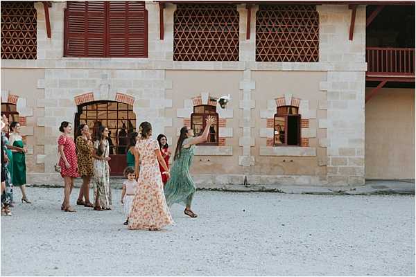 The bouquet toss is taking place outdoors in a gravel courtyard in front of a large stone building with dark red wooden shutters and decorative lattice window grilles. Approximately eight to ten female guests are gathered, with one woman in a sage green tiered midi dress reaching up to catch the bouquet mid-air, while the bride in a floral pink and white maxi dress has her back turned after throwing it. The guests are dressed in a variety of colorful outfits including emerald green, red floral, and mixed-print dresses, suggesting a relaxed, warm-weather reception. The shot is a wide candid frame capturing the full motion of the toss against the chateau-style facade.