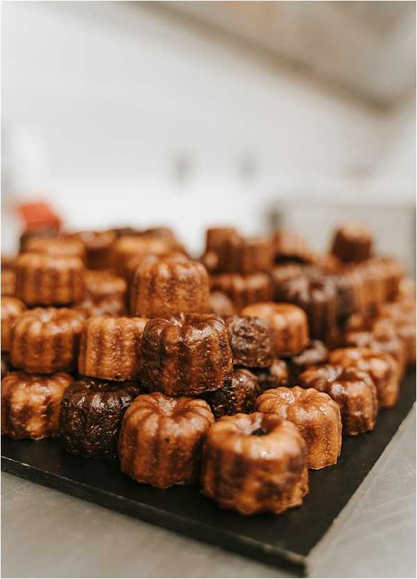 Close-up detail shot of a large pile of canelés — traditional French Bordeaux pastries — displayed on a dark slate board at what appears to be a wedding reception or cocktail hour. The canelés range in color from golden amber to deep dark brown, indicating varying degrees of caramelization on their fluted exteriors. Approximately 30 or more individual pastries are stacked and arranged in a generous mound. The background is softly blurred, showing a light-colored interior setting.